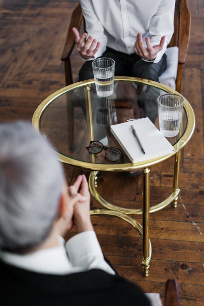 Two adults discussing mental health in a counseling session across a glass table indoors.